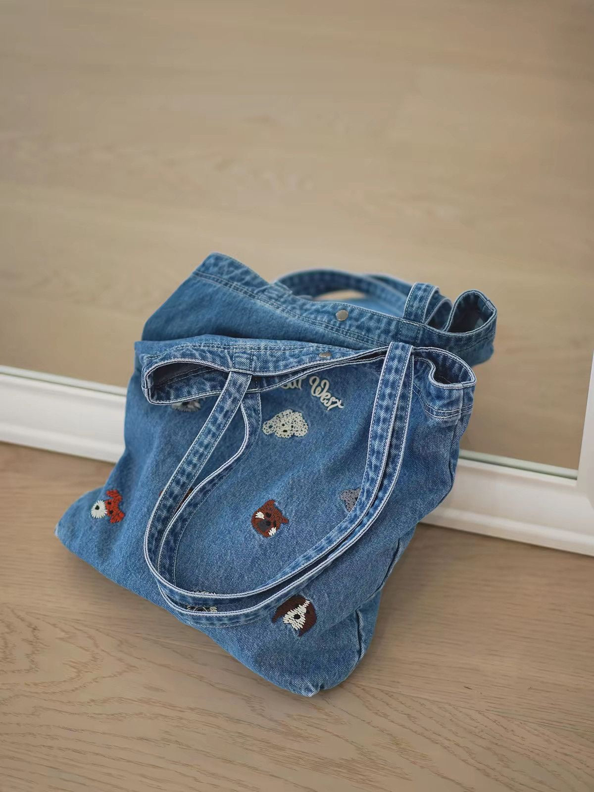 Washed-blue denim tote with embroidered animal motifs styled on a wooden floor